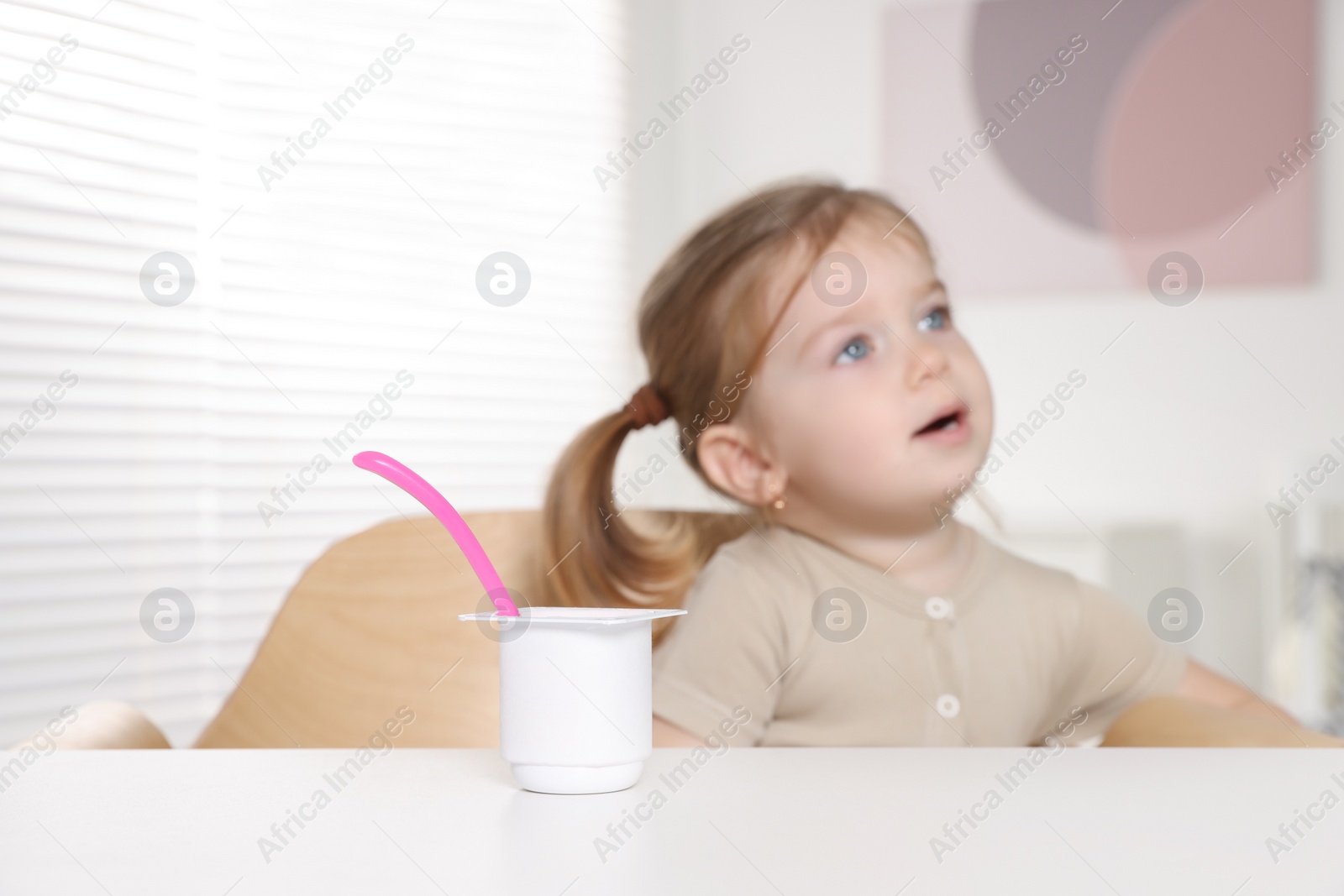 Photo of Cute little child eating tasty yogurt from plastic cup with spoon at white table indoors