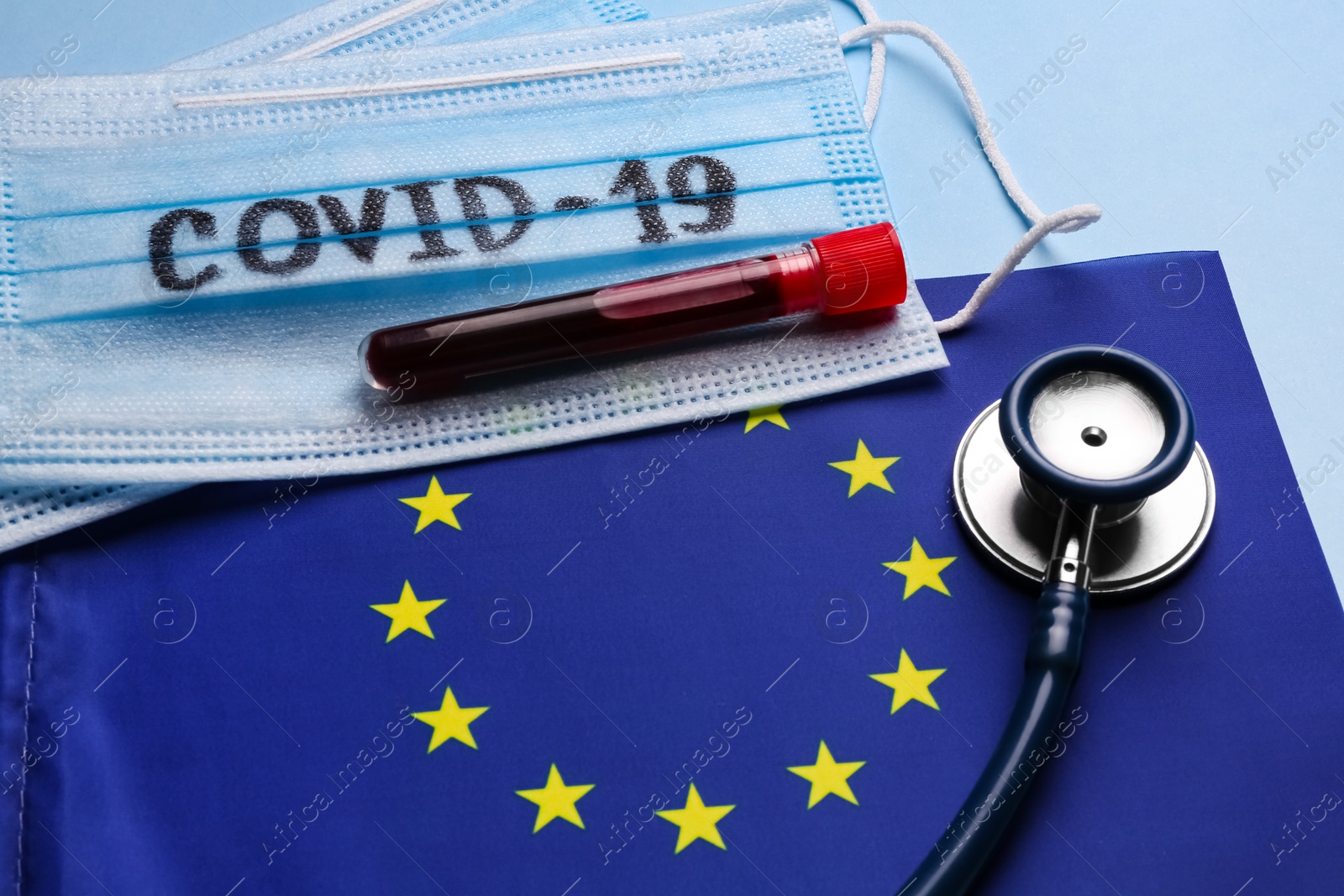 Photo of European Union flag, protective masks and test tube with blood sample on light blue background. Coronavirus outbreak