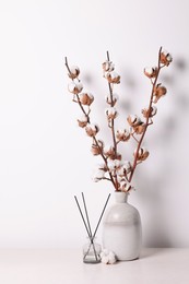 Photo of Reed diffuser and vase with cotton branches on table against white background