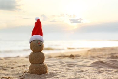 Photo of Snowman made of sand with Santa hat on beach near sea at sunset, space for text. Christmas vacation