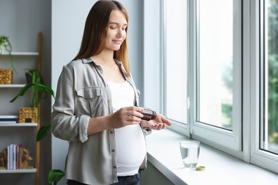 Photo of Beautiful pregnant woman taking pill from bottle near window at home