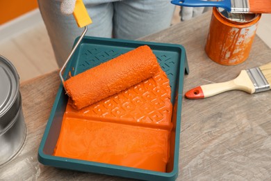 Photo of Woman taking orange paint with roller from tray at table, closeup