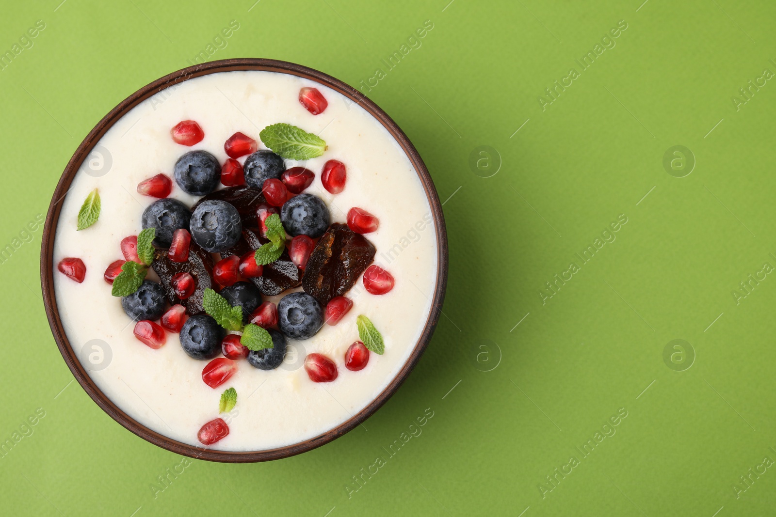 Photo of Delicious semolina pudding with blueberries, pomegranate, dates and mint in bowl on green table, top view. Space for text