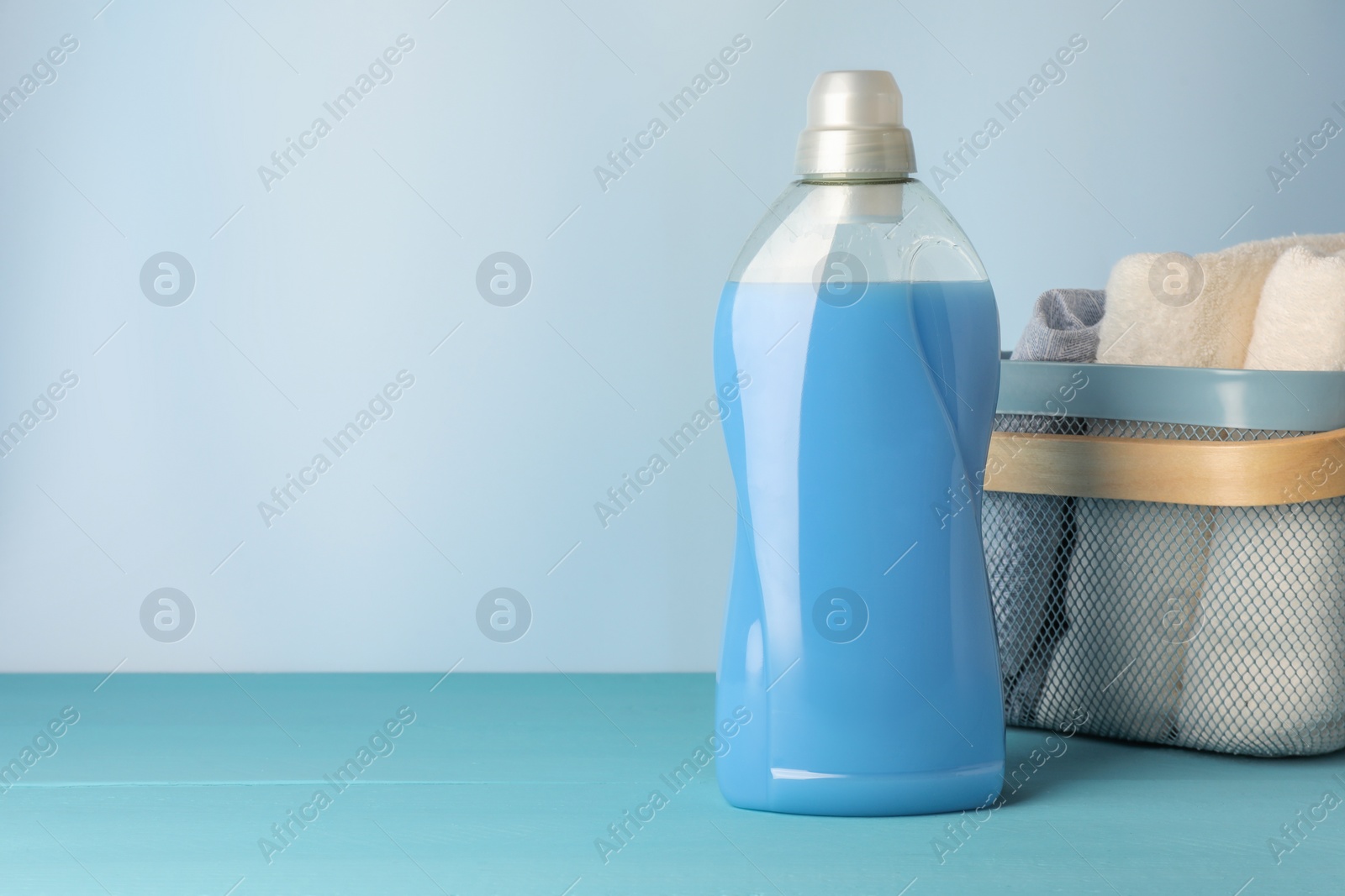 Photo of Bottle of fabric softener and towels in basket on light blue table, space for text