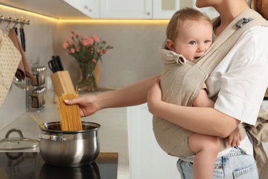 Mother holding her child in sling (baby carrier) while cooking pasta in kitchen