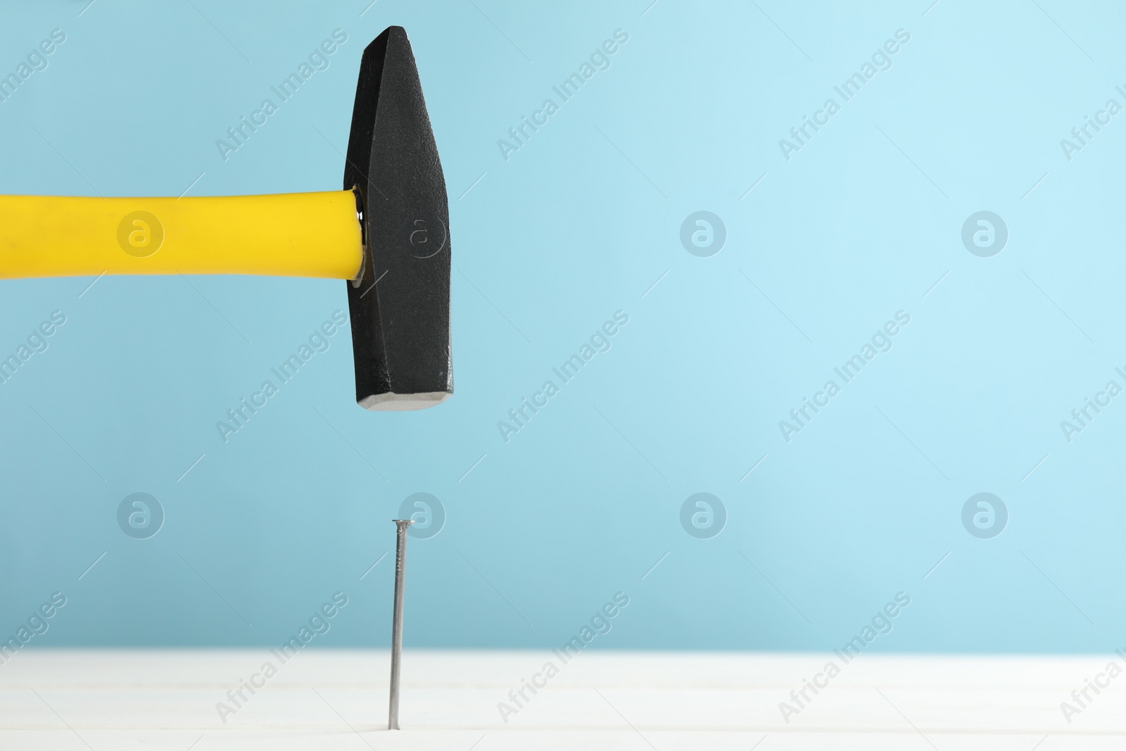 Photo of Hammering metal nail on white wooden table against light blue background. Space for text