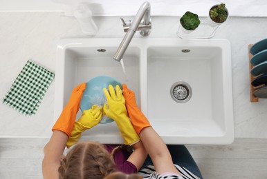 Photo of Mother and daughter in protective gloves washing plate above sink, top view