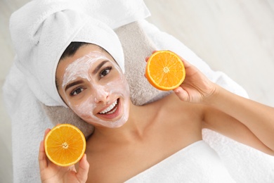 Pretty woman with rejuvenating facial mask holding sliced orange in spa salon, above view