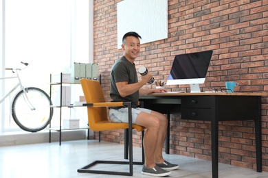 Photo of Young man lifting weights and using computer in office. Workplace fitness