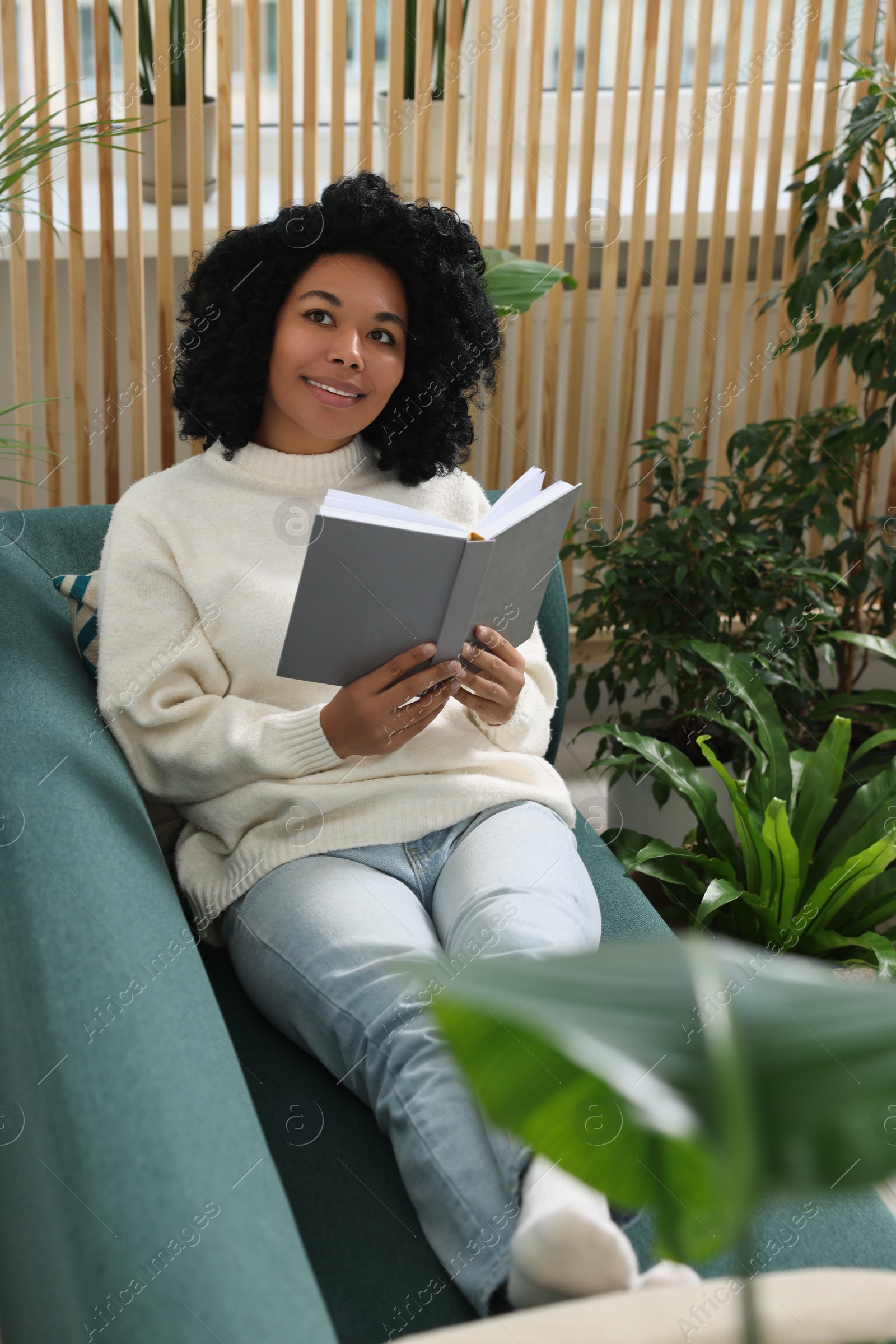 Photo of Relaxing atmosphere. Happy woman with book on sofa near beautiful houseplants in room