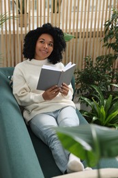 Relaxing atmosphere. Happy woman with book on sofa near beautiful houseplants in room