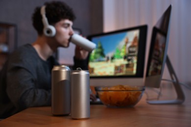 Young man with energy drinks playing video game at wooden desk indoors, focus on cans