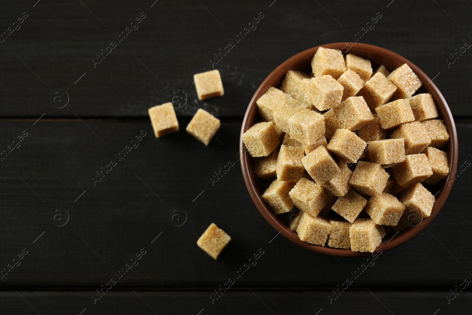 Photo of Brown sugar cubes in bowl on black wooden table, top view. Space for text