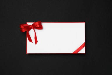 Photo of Blank gift card with red bow on black background, top view
