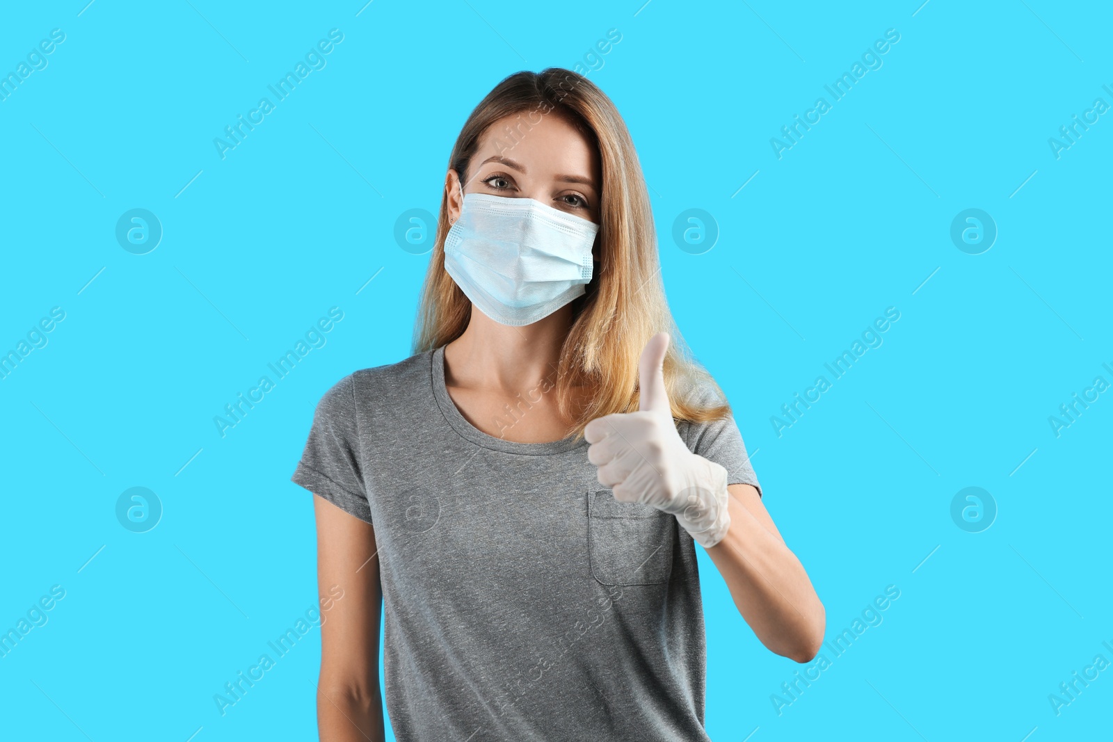 Photo of Young woman in medical gloves and protective mask showing thumb up on light blue background
