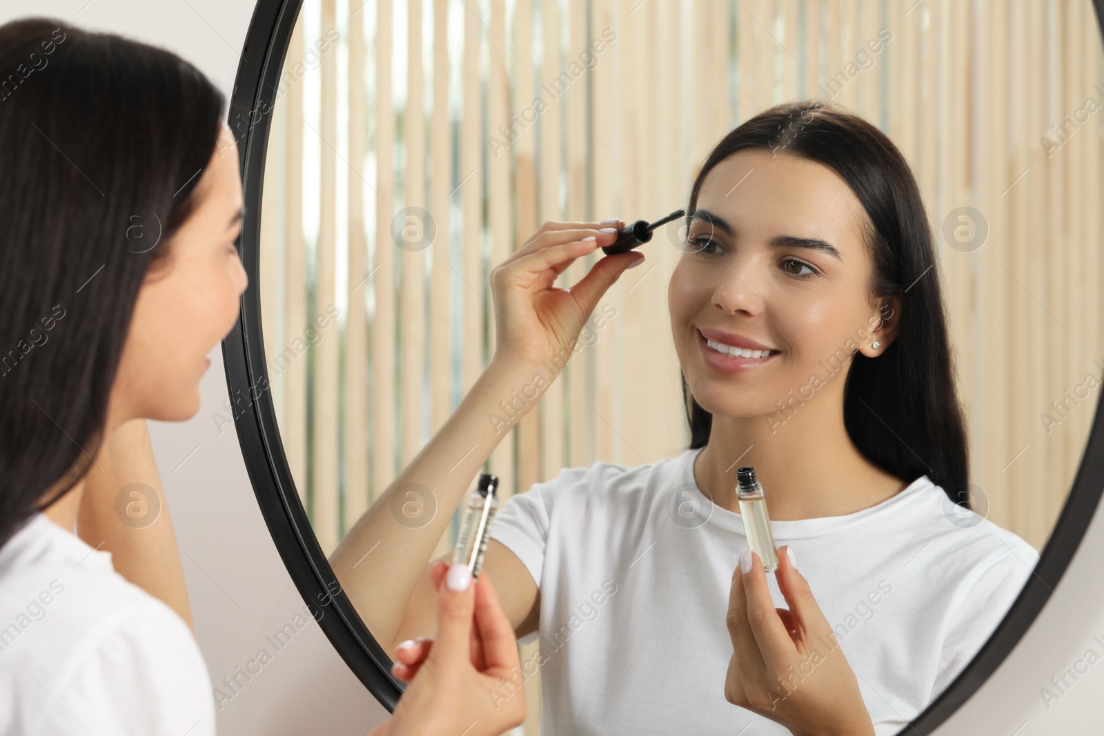 Photo of Young woman applying oil onto eyelashes near mirror indoors