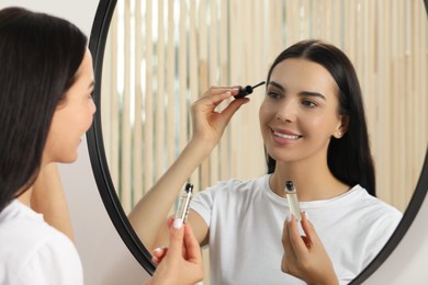 Young woman applying oil onto eyelashes near mirror indoors