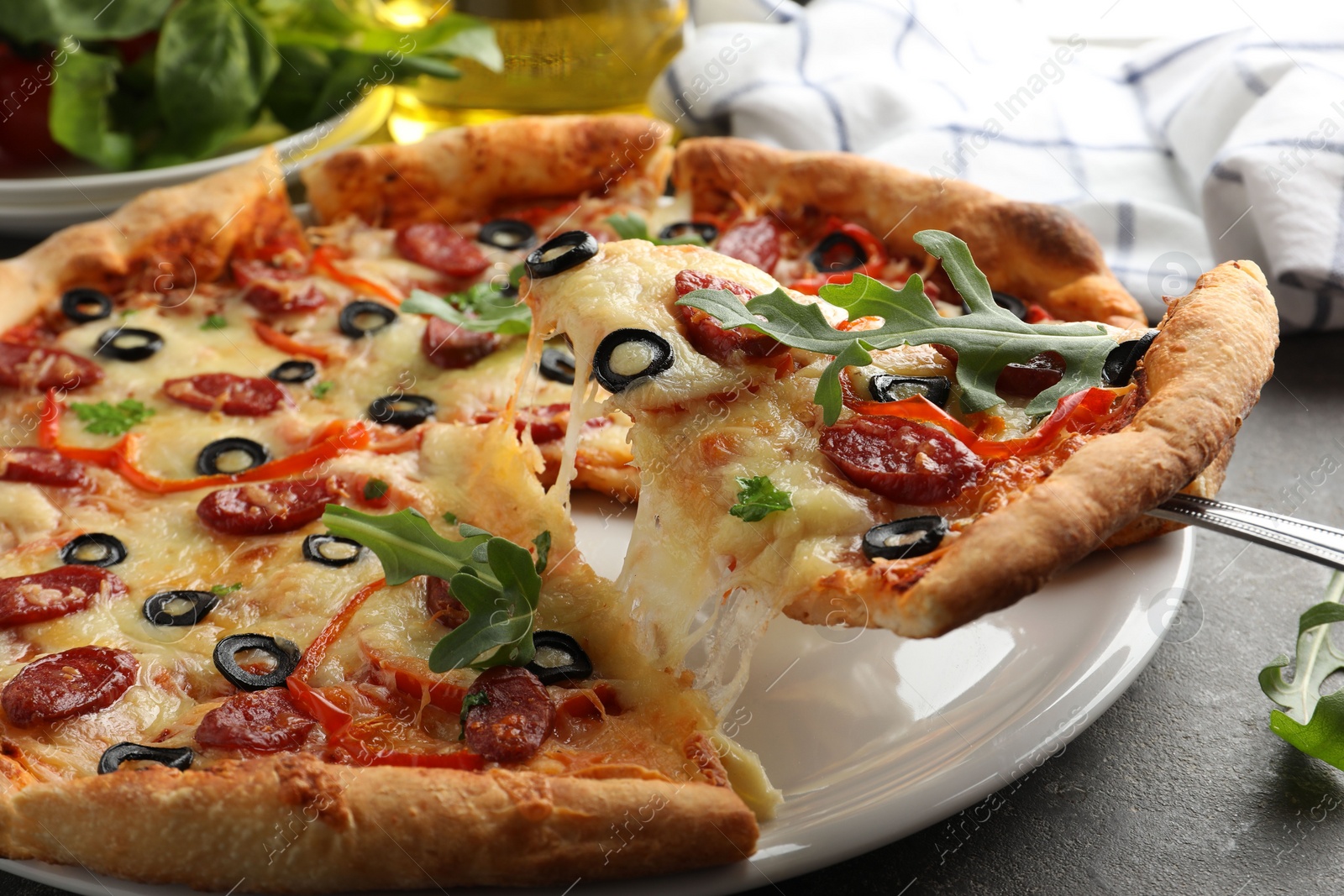 Photo of Taking piece of tasty pizza with smoked sausages, olives, pepper and parsley at gray table, closeup