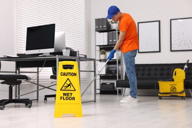 Photo of Cleaning service worker washing floor with mop, focus on wet floor sign in office