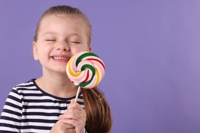 Photo of Happy little girl with colorful lollipop swirl on violet background, space for text