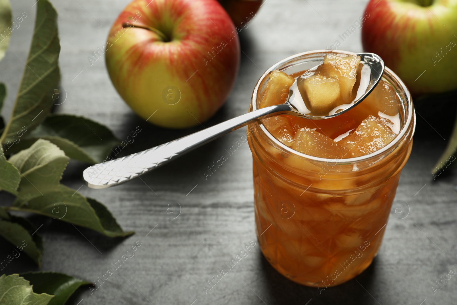 Photo of Tasty apple jam with spoon in glass jar and fresh fruits on grey table