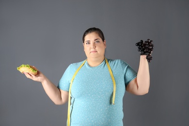 Photo of Overweight woman with hamburger, grapes and measuring tape on gray background