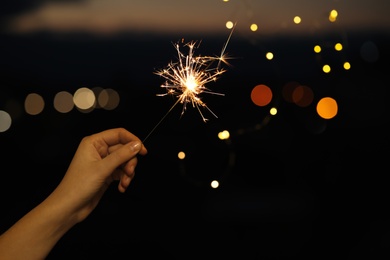 Photo of Woman holding bright sparkler against blurred lights, closeup