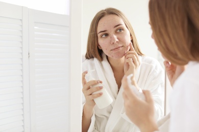 Young woman with acne problem holding bottle with cosmetic product in bathroom