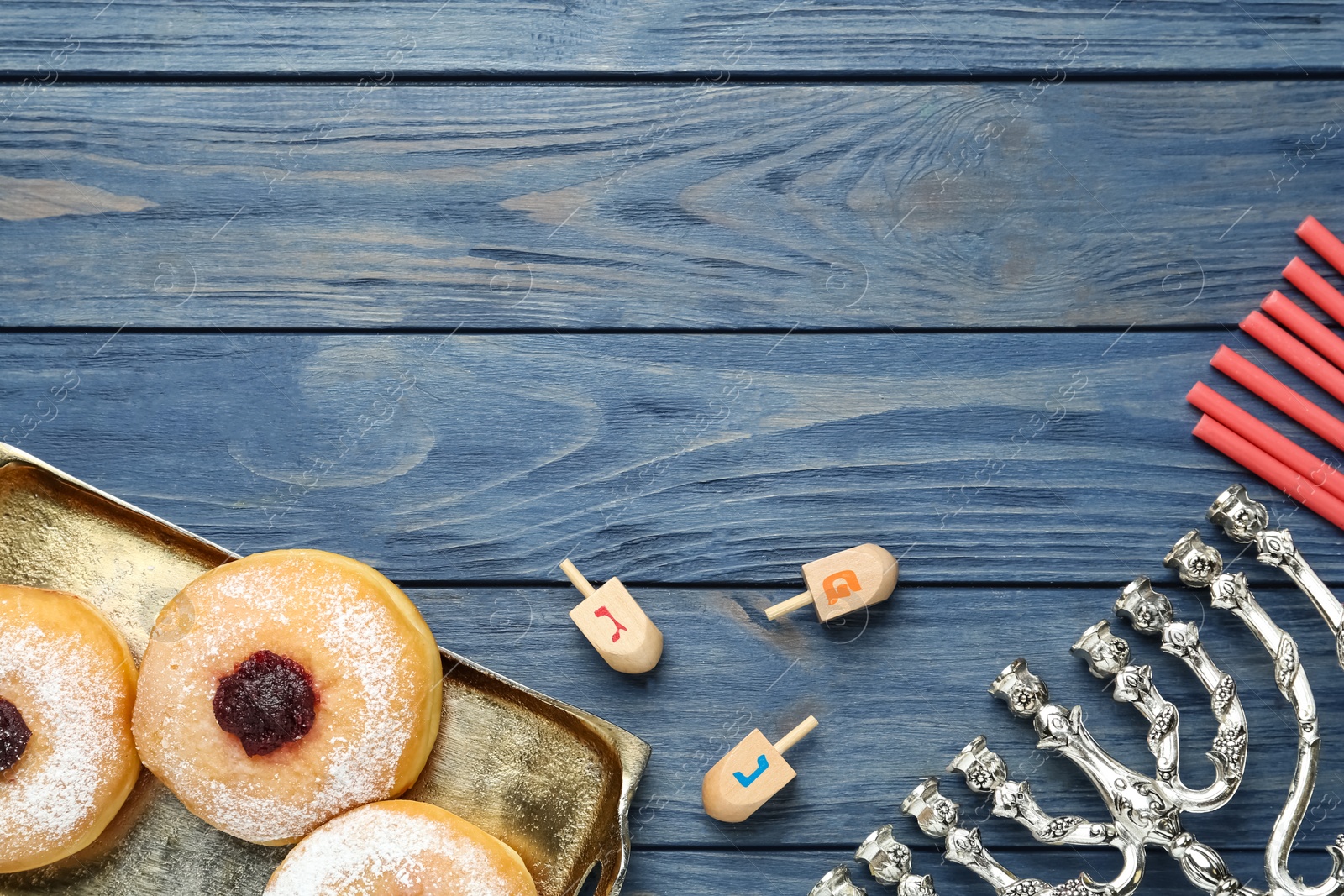 Photo of Flat lay composition of menorah, red candles, dreidels with He, Pe, Nun, Gimel letters and sufganiyot on blue wooden table, space for text. Hanukkah symbols