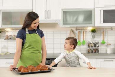 Photo of Son and mother with tray of oven baked buns at table in kitchen