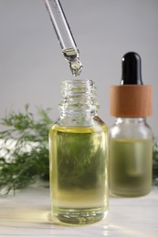 Photo of Dripping dill essential oil from pipette into bottle on white table, closeup