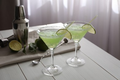 Photo of Delicious Margarita cocktail in glasses, limes and bartender equipment on white wooden table