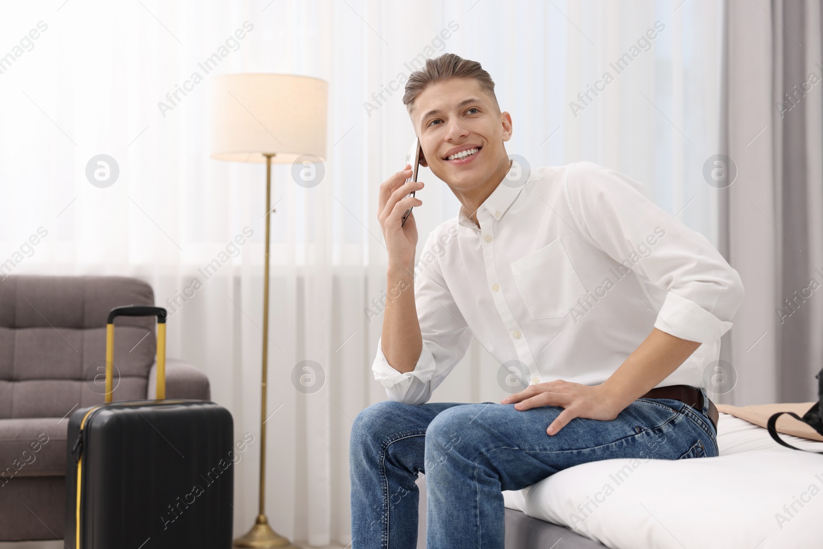 Photo of Smiling guest talking by smartphone on bed in stylish hotel room