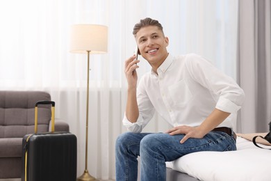 Photo of Smiling guest talking by smartphone on bed in stylish hotel room