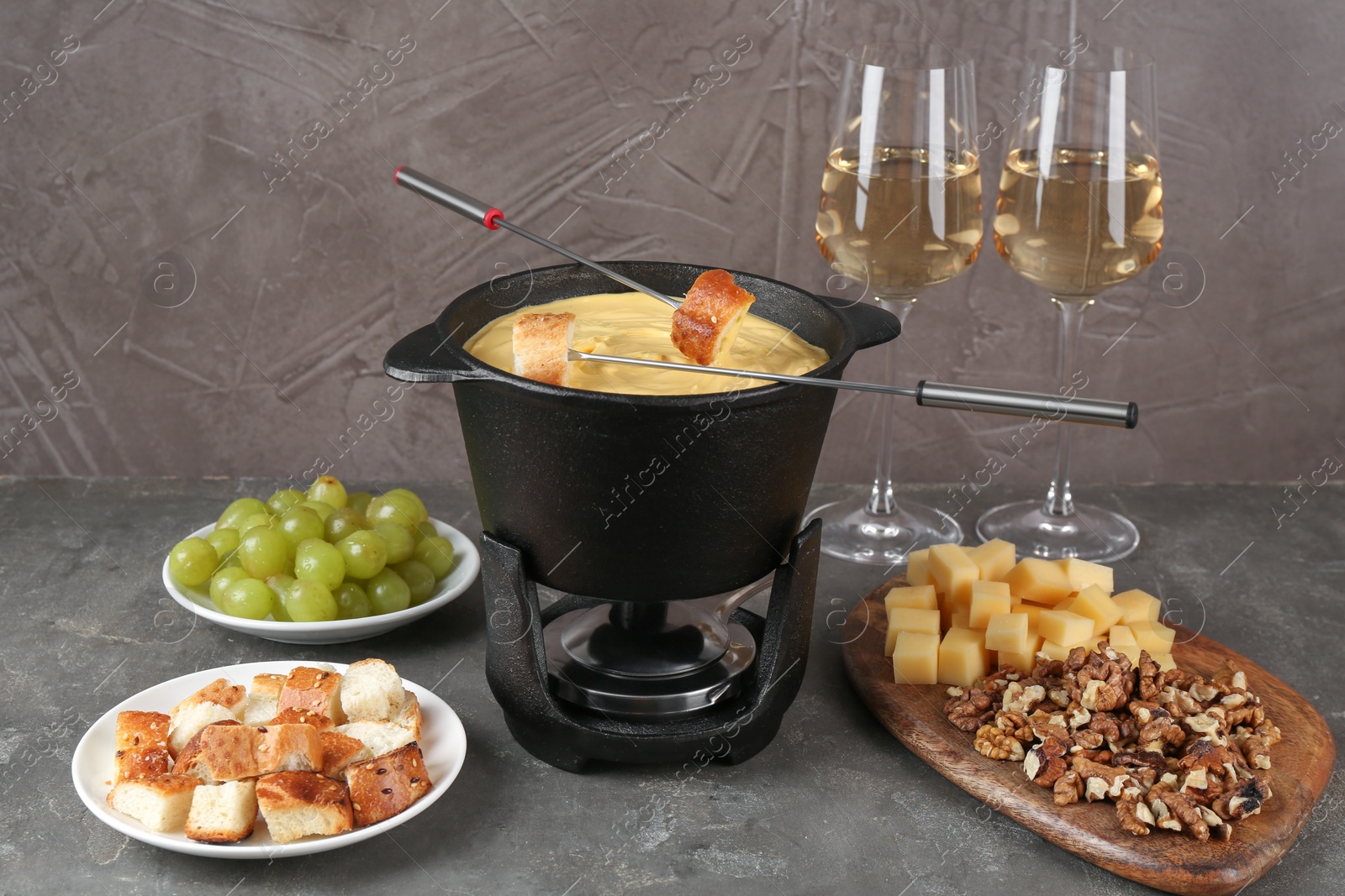 Photo of Fondue pot with tasty melted cheese, forks, wine and different snacks on grey table