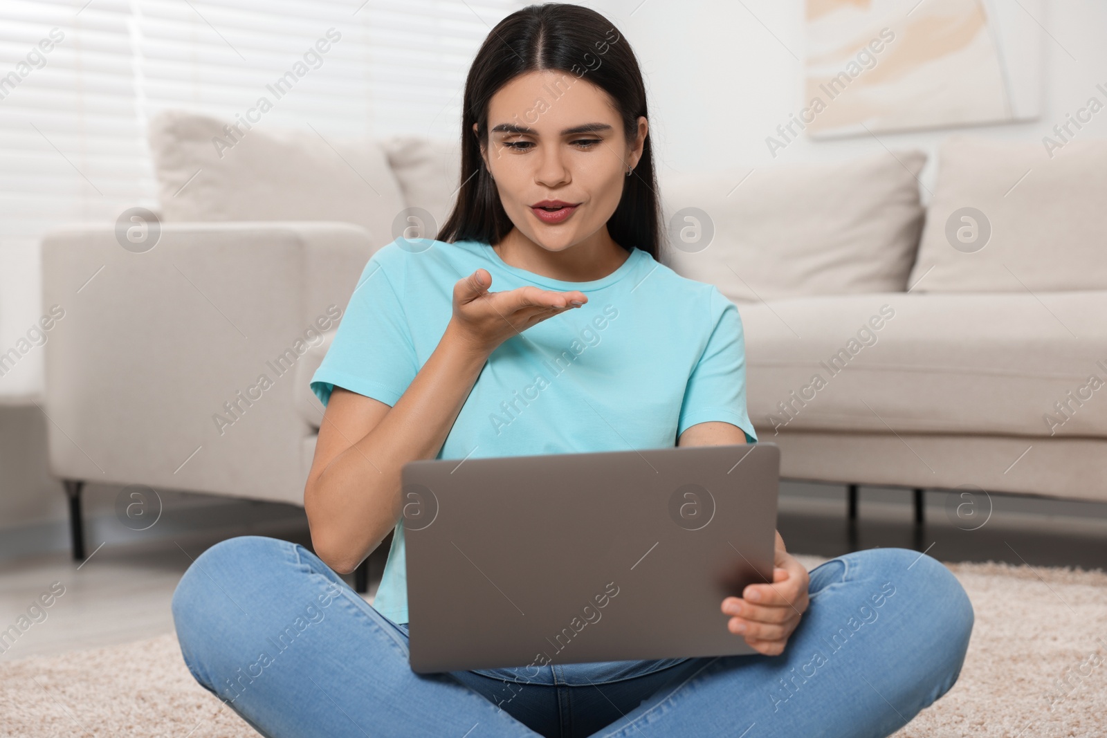 Photo of Young woman having video chat via laptop and blowing kiss on floor at home