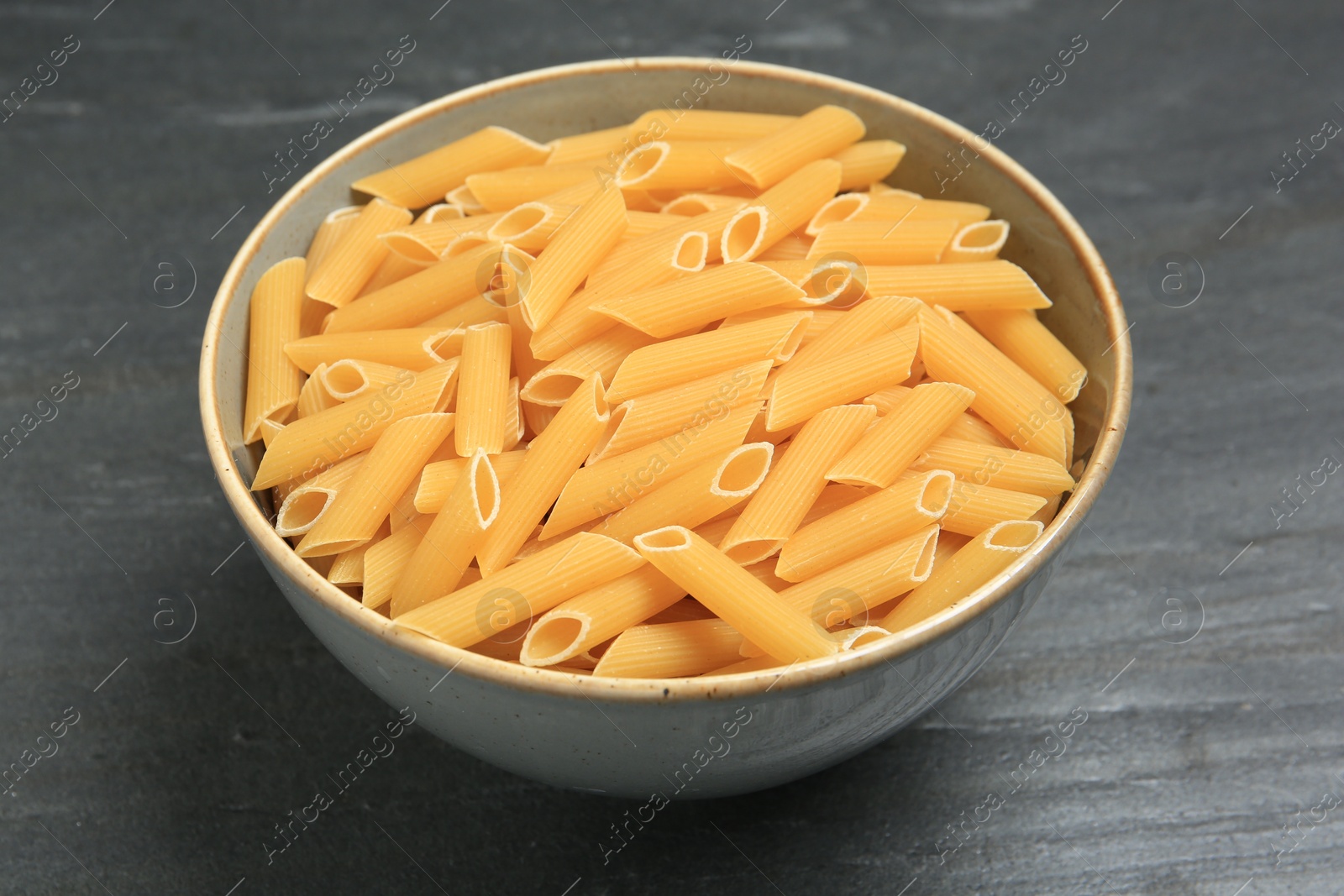 Photo of Raw penne pasta in bowl on grey table, closeup