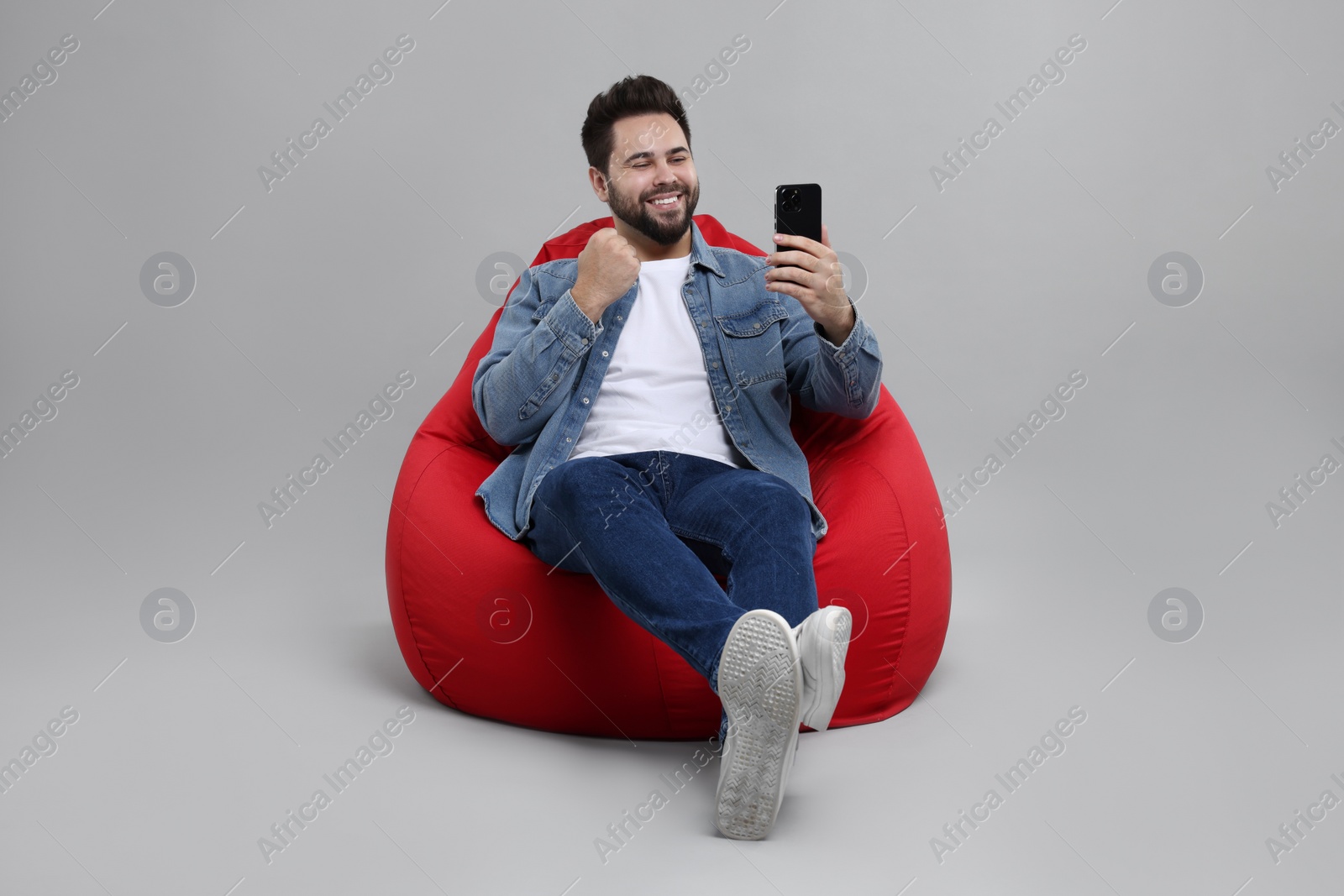 Photo of Happy young man using smartphone on bean bag chair against grey background