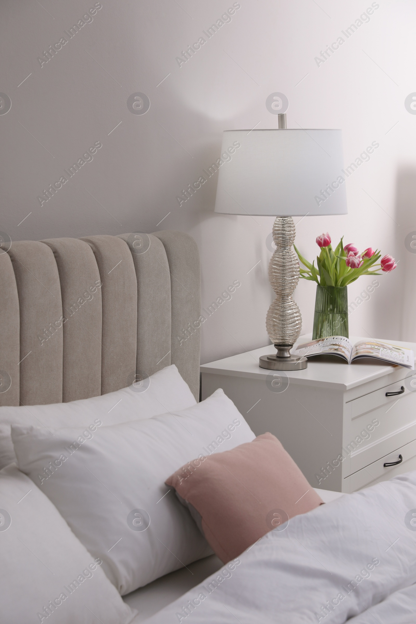 Photo of Stylish lamp, flowers and magazine on bedside table indoors. Bedroom interior elements