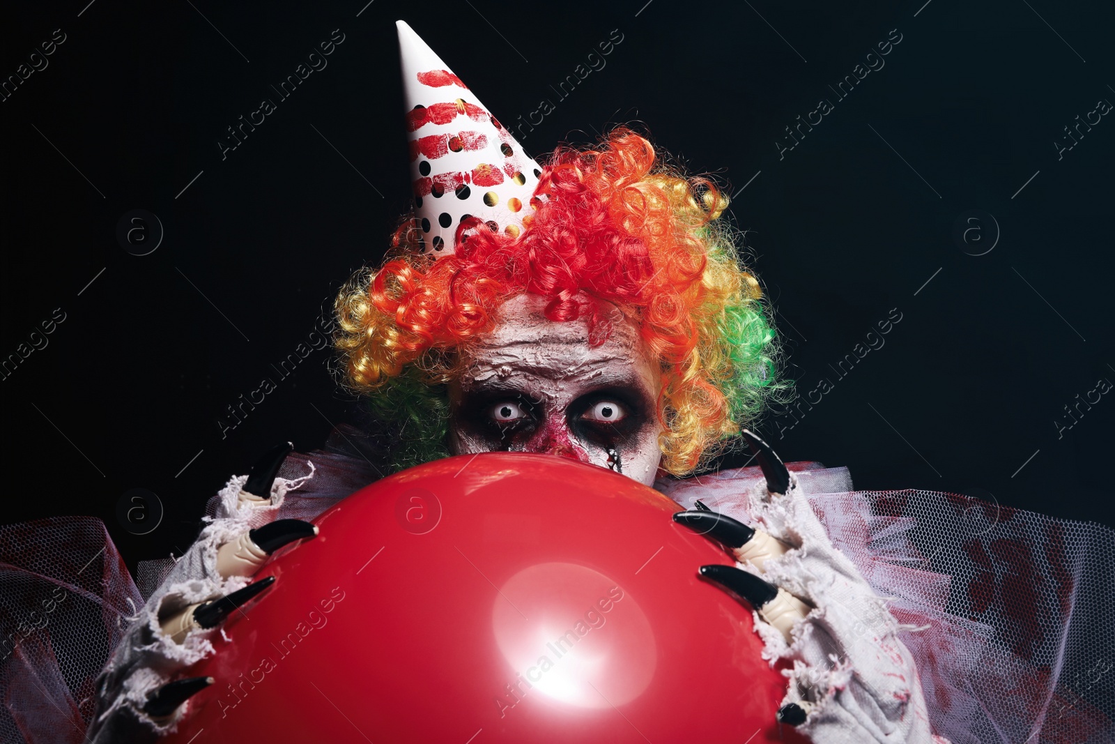 Photo of Terrifying clown with red air balloon on black background. Halloween party costume