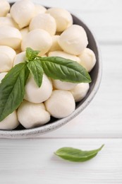 Photo of Tasty mozzarella balls and basil leaves in bowl on white wooden table, closeup