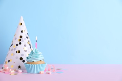 Delicious birthday cupcake with burning candle, marshmallows and party decor on pink table against light blue background, space for text