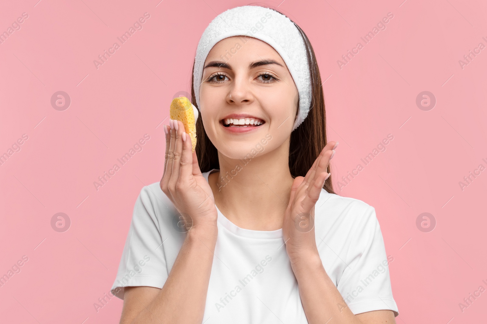 Photo of Young woman with headband washing her face using sponge on pink background