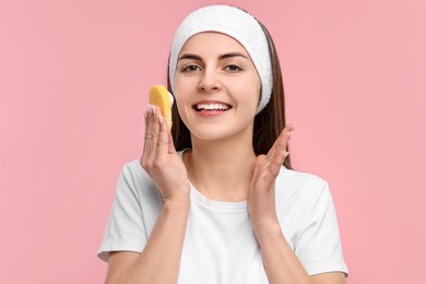 Photo of Young woman with headband washing her face using sponge on pink background