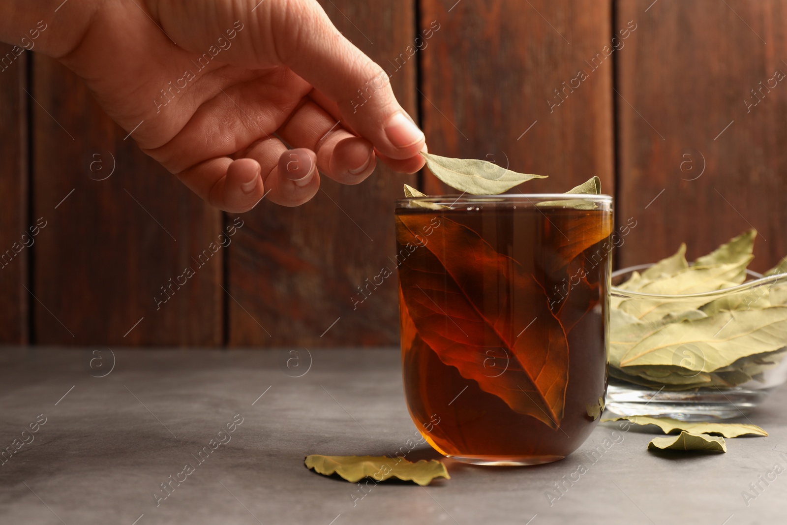 Photo of Woman putting bay leaf into cup of freshly brewed tea at grey table, closeup