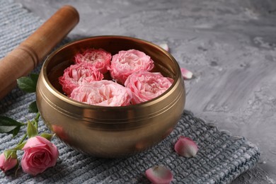 Photo of Tibetan singing bowl with water, beautiful roses and mallet on grey textured table, closeup. Space for text