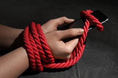 Woman with roped hands holding smart phone on grey background, closeup. Solitude concept