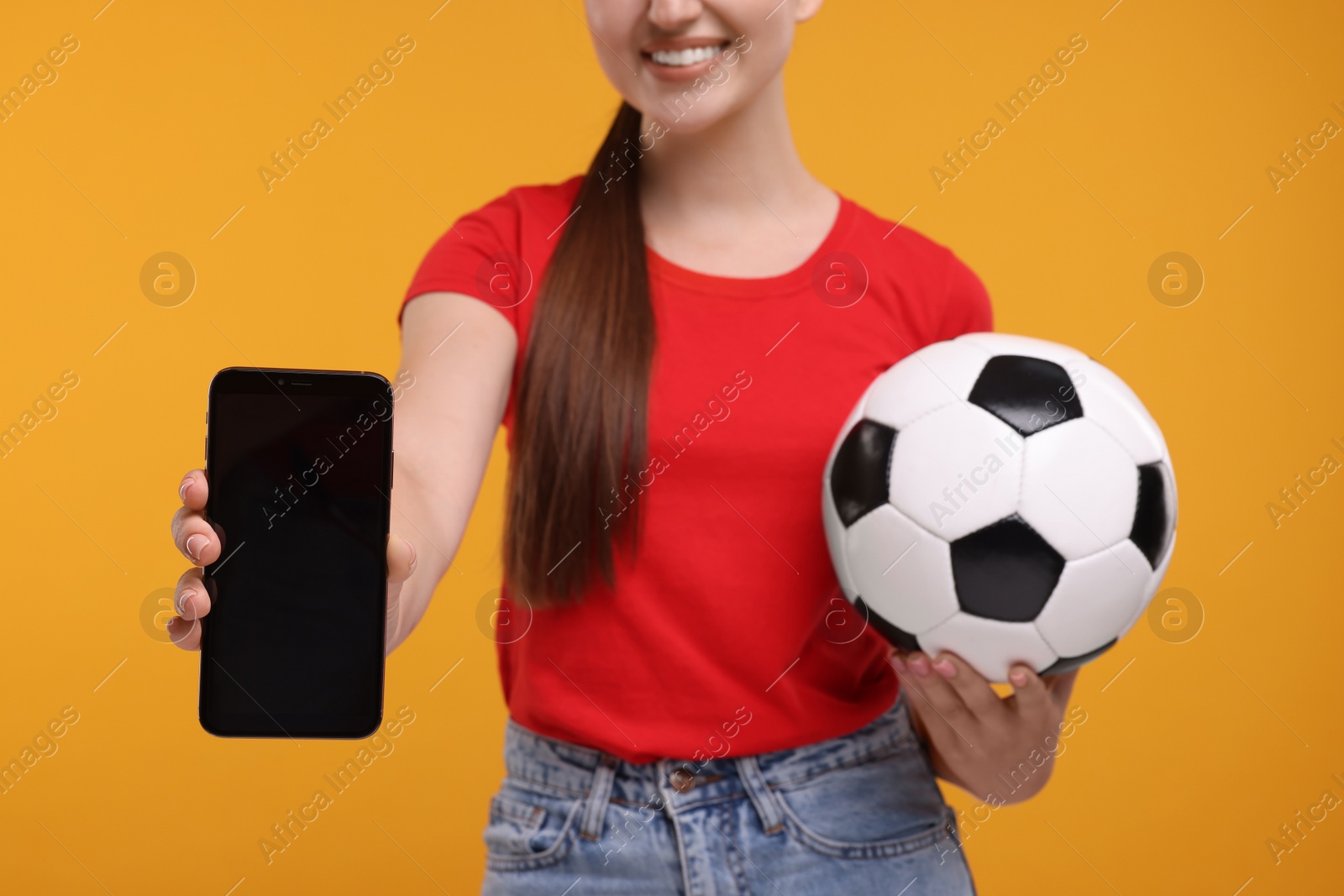 Photo of Happy soccer fan with ball showing smartphone on orange background, closeup