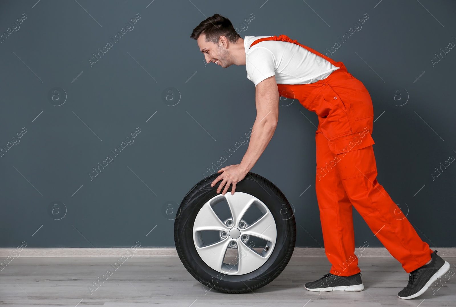 Photo of Young mechanic in uniform with car tire near dark wall