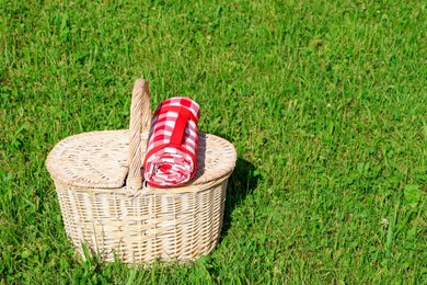 Photo of Rolled checkered tablecloth with picnic basket on green grass outdoors, space for text